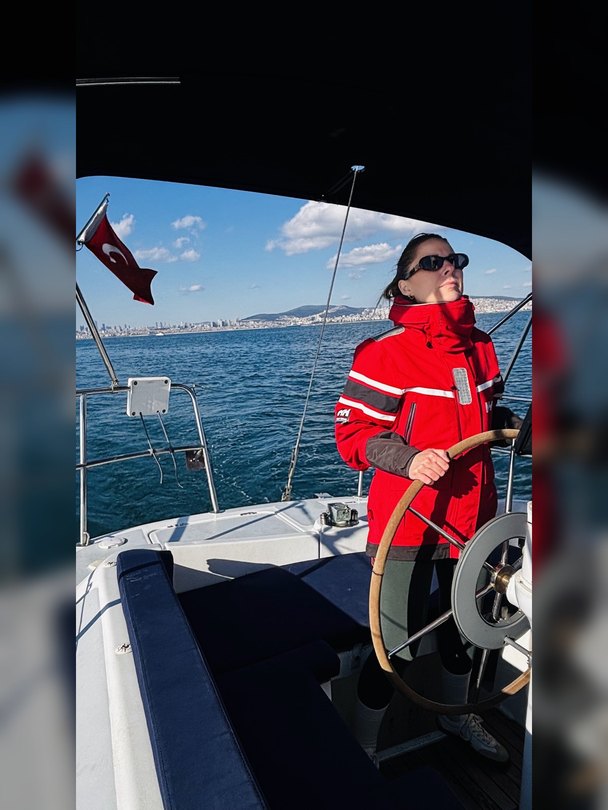 Dümen başında kırmızı montlu katılımcı ve İstanbul kıyısı